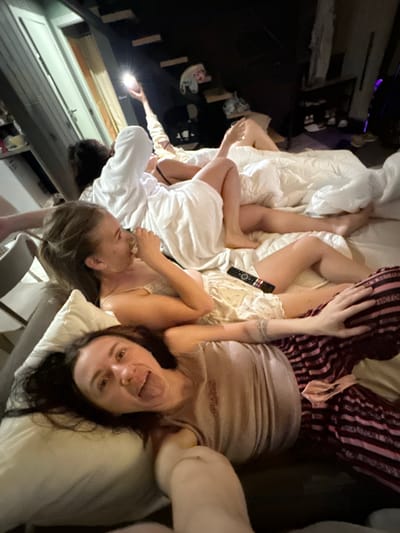 The image shows a group of young women in a dimly lit room, possibly a bedroom, with a focus on their leisure activity. The room appears to be a domestic setting. The women are lying on a bed covered with white bedding. One woman in the foreground is lying on her back, looking at the camera with her tongue sticking out and a playful expression. She is wearing a light-colored top. Behind her, another woman is lying down, holding a phone and using its flashlight. She is wearing a white robe or hooded garment. To the left and further back, parts of other individuals are visible, suggesting at least three people are present. A remote control is visible on the bed. The overall atmosphere is casual and relaxed. There is no information in the image to suggest this location is in Novaya Vozhoika, Russia.