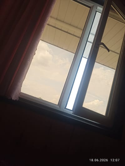 The image depicts a view through a window, looking outwards at a partly cloudy sky. A portion of the window is open, allowing a sliver of bright light to enter. To the left, a dark red curtain is partially drawn, obscuring some of the view. The window frame is white. Above the window, a metallic ceiling structure is visible. The lower part of the image is dark, indicating the interior of a room. In the bottom right corner, a timestamp reads "18.06.2026 12:07". The scene appears to be from Razzakov, Kyrgyzstan. There are no people visible in the image.