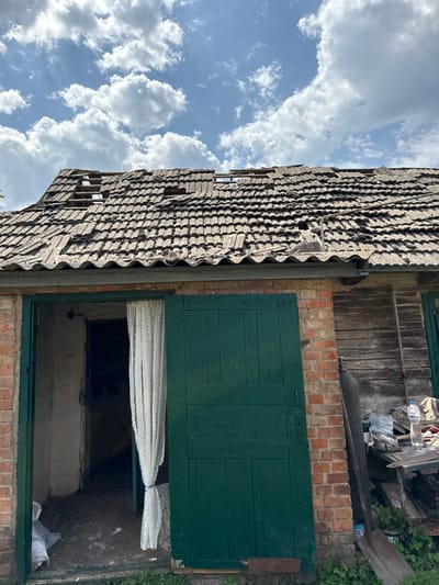 The image depicts the exterior of a building in Nedryhailiv, Ukraine, under a bright blue sky with scattered white cumulus clouds. The building features brick construction on the left and vertical wooden planks on the right side of the facade. The most prominent feature is the severely damaged roof, composed of displaced and missing clay tiles, revealing the underlying wooden support structure in multiple areas. A dark, corrugated eave runs along the lower edge of the roof.

On the left, an open doorway reveals a dark interior space with a dirt floor and rough, unpainted walls. A white, lace-patterned curtain is tied back, hanging vertically within this opening. To the right of the open doorway, a large, dark green wooden door, appearing to be a single leaf of a double door, is partially open or slightly ajar. Further to the right, a section of wooden planking forms part of the wall, alongside which a tall, dark cylindrical object, possibly a gas tank, leans. A long-handled shovel with a wide blade is propped against the wall nearby. On the ground to the right, a collection of wooden planks forms a makeshift surface, upon which two plastic water bottles, one full and one partially empty, are visible among other unidentifiable items. Green vegetation is present at the base of the building. No individuals are visible in the scene.