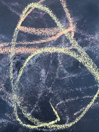 The image is a close-up, high-angle shot of a dark surface, likely a chalkboard or dark paper, with colorful chalk markings. The chalk lines are primarily in shades of yellow, pinkish-orange, and some faint purple and white. The markings appear to be abstract scribbles and shapes, with some thicker, more defined lines forming loops and arcs, and thinner, fainter lines creating a scattered effect. The chalk has a textured, powdery appearance, and there are areas where the chalk dust is more concentrated, giving the impression of a slightly smudged or layered effect. The background is a uniform dark color, showing the texture of the surface. There are no people or recognizable objects in the image. The image does not contain any information that can be used to determine its location within the city of Novoterksky, Russia.