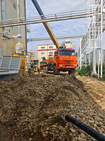 An orange Kamaz truck with a crane boom is parked on a construction site. The truck is positioned on a dirt and gravel surface in front of a large industrial building. In the background, there is a yellow construction ladder and a white building with multiple windows. A person is visible in the driver's seat of the truck, and another person is standing near the crane. To the left of the truck, a yellow construction lift is visible. The ground in the foreground is covered in a large pile of rocks and dirt. The sky above is partly cloudy. The location is identified as Konokovo, Russia.
