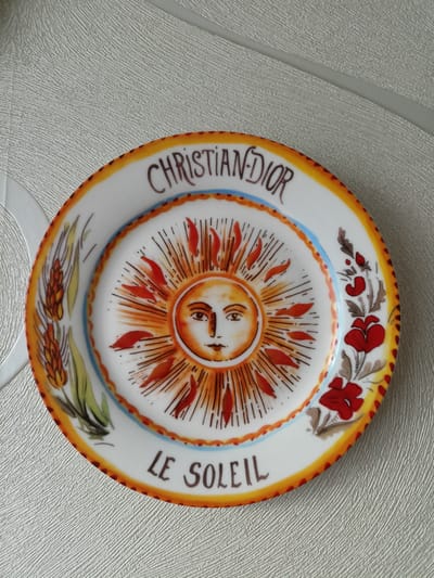 This is a close-up, overhead shot of a decorative ceramic plate. The plate has a white background and is adorned with a colorful illustration of a sun with a human-like face at its center. The sun's rays are depicted in orange and red, emanating outwards. Around the sun, there are stylized depictions of wheat stalks on the left side and red flowers with brown accents on the right. The rim of the plate is decorated with a yellow and orange border.
Visible text on the plate reads "CHRISTIAN DIOR" in dark brown lettering arched across the top, and "LE SOLEIL" in similar lettering arched across the bottom. The overall style of the illustration is somewhat folk art or naive.
The plate is placed on a textured surface that appears to be a light-colored tablecloth or placemat with a raised, ribbed pattern. The lighting is bright and even, suggesting it might be daytime, but there are no specific clues about the time of day. The setting appears to be indoors, on a table. The image does not depict any people or a specific event, but rather focuses on the object itself. The location context provided is Volgodonsk, Russia, but this is not visually evident from the image.