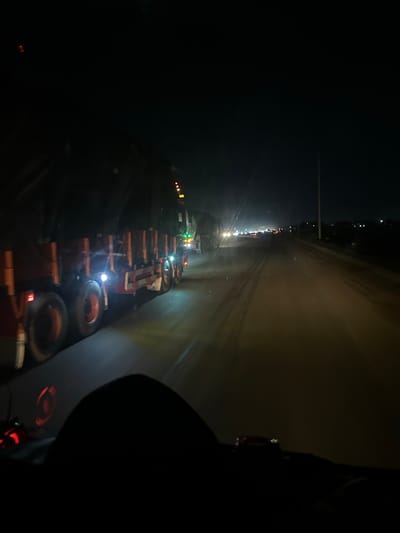 The image depicts a nighttime scene on a highway, likely in or near Ibafo, Nigeria. Multiple large trucks are seen traveling in the same direction, with their headlights and taillights illuminating the dark road. The truck in the foreground is a flatbed truck, partially covered with a dark tarp, and its wheels and side are visible. The road appears to be a dirt or unpaved surface, with visible tire tracks. In the distance, more vehicles are visible, suggesting a traffic jam or slow-moving traffic. The overall impression is one of a busy and active transportation route at night.