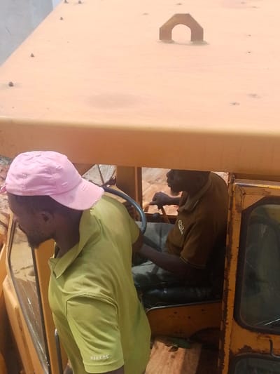 Two adult males are observed in proximity to and within the cabin of a heavy, yellow-orange piece of machinery in an outdoor, dirt-floored environment in the city of Agukwu Nri, Nigeria. The male on the left, partially obscured by the machinery's roof, wears a distressed pink baseball cap and a lime green short-sleeved polo shirt with "MAKERS" embroidered on the right sleeve. He has a beard and is positioned with his back mostly to the viewer, looking towards the right. The second male is seated inside the machinery's cabin, wearing a dark brown or olive green short-sleeved polo shirt featuring a light-colored logo on the chest. He also has a beard and is actively engaged with the machinery's controls, with his hands on the steering wheel and a lever. The machinery's cabin window on the right exhibits visible cracking. The immediate background shows reddish-brown earth.