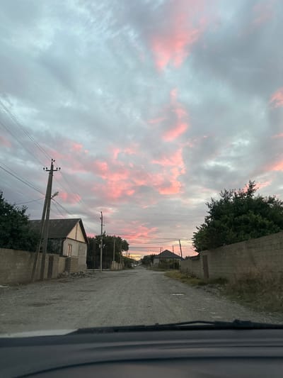 The image depicts a rural scene in Miatli, Russia, likely during sunset or sunrise, indicated by the soft pink hues in the overcast sky. The perspective is from within a vehicle, looking out towards a dirt road that stretches into the distance. On the right side of the road, a grey brick wall runs along the edge of a property, with a large tree providing shade. Further down the road, there are several houses with peaked roofs and visible windows, some illuminated from within. Telephone poles with wires crisscross the scene, adding to the rural atmosphere. The overall mood is tranquil and serene, capturing a quiet moment in a village setting. There is no visible text in the image.