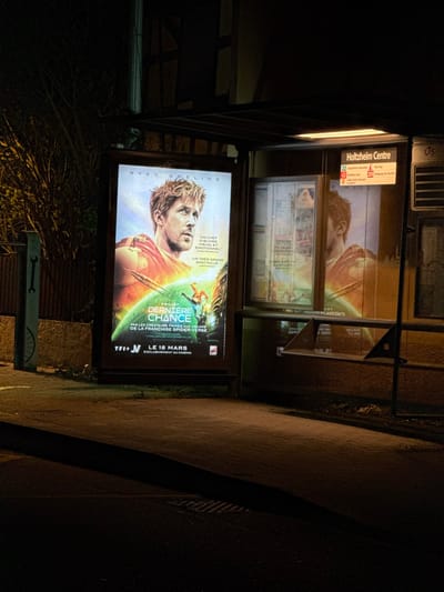 The image depicts a nighttime urban scene at a bus stop in Holtzheim, France. The central feature is an illuminated advertising billboard displaying a movie poster for "PROJET DERNIERE CHANCE" (Project Last Chance), starring Ryan Gosling. Gosling, with blonde hair, is depicted wearing a red and orange garment against a light blue and white sky. Below him, a green, wave-like structure with a figure surfing is visible. French text on the poster mentions "UN CHEF D'OEUVRE VISUEL ET ÉMOTIONNEL", "UN TRÈS GRAND SPECTACLE", "PAR LES CRÉATEURS PRIMÉS AUX OSCARS DE LA FRANCHISE SPIDER-VERSE", "TFI+ J", and "LE 18 MARS EXCLUSIVEMENT AU CINEMA".

Adjacent to the billboard is a bus stop shelter with a dark metal frame and glass panels. An overhead light illuminates the interior of the shelter. A white sign prominently displays "Holtzheim Centre" at the top, followed by bus line numbers (22, 40, 42, 209) and their respective destinations. The movie poster is reflected in the bus stop's glass panels. A dark, low bench-like structure is visible inside the shelter. The ground in the foreground is a dark, paved surface, partially illuminated by the light from the billboard and bus stop. To the left of the billboard, a dark utility pole or similar structure with a white wrench symbol is partially visible. The background consists of dark foliage and indistinct building structures, further emphasizing the nighttime setting. No individuals are present in the scene.