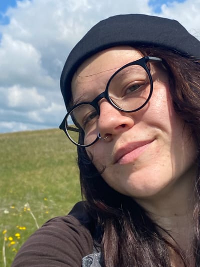 A close-up, selfie-style shot shows a person wearing a black beret and black-rimmed glasses. They have a nose ring and long, dark brown hair. The background is a grassy, rolling hill under a partly cloudy blue sky, suggesting an outdoor, rural or park setting. The lighting indicates it is daytime, possibly sunny. The provided location of Petrovskoye, Russia, is not discernible from the visual information in the image itself.