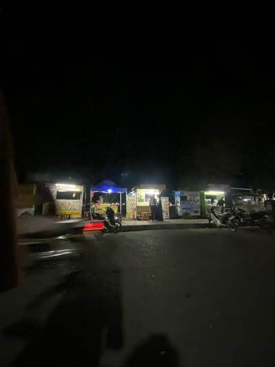 The image depicts a nighttime scene at a street food stall area in Tanggul, Indonesia. Several food stalls are visible, illuminated by artificial lights, with various food items and drinks displayed. Some stalls have awnings or tents, with one particularly prominent blue tent. Several motorcycles are parked in front of the stalls, and some individuals appear to be working or patronizing the stalls. The ground in front of the stalls is wet, reflecting the lights, suggesting recent rain or cleaning. The overall environment is that of an active, albeit dimly lit, street market.