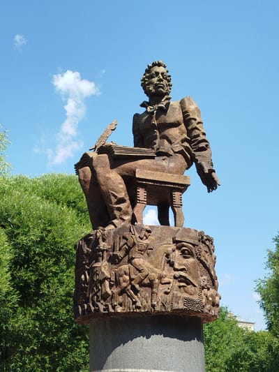 A bronze statue of a seated man with a quill pen and a book is displayed outdoors. The man has curly hair and is dressed in a jacket and shirt. The base of the statue is decorated with intricate reliefs of people and animals. The statue is located in Yalutorovsk, Russia, against a backdrop of lush green trees and a clear blue sky with a few white clouds. The statue is positioned on a stone pedestal.