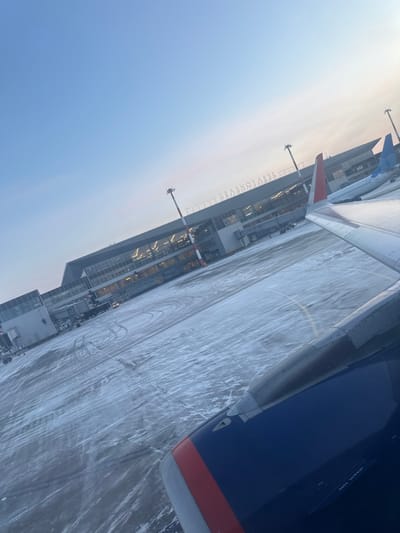 A ground-level view from an aircraft window at Yemelyanovo Airport, Krasnoyarsk, Russia, shows the airport tarmac and terminal building under a clear, pale blue sky. The foreground features the dark blue engine nacelle with a red stripe and a portion of the white wing of the viewing aircraft. The tarmac is covered with a thin layer of snow or ice, exhibiting numerous tire tracks. In the midground, a modern, multi-story terminal building with large glass windows is visible, illuminated from within. The word "KRASNOYARSK" is displayed on its roofline. A jet bridge extends from the terminal towards a gate position, with a ground support vehicle partially visible near its base. Several tall, red-and-white striped light poles stand on the tarmac. Two additional commercial aircraft, predominantly white with blue tail liveries, are parked near the terminal building further in the background. No discernible individuals are visible, but the illuminated terminal and operational elements suggest human presence and activity.