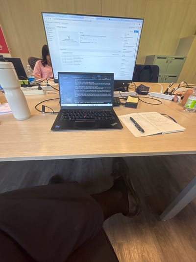 The image depicts a desk in an office environment, likely in Basiglio, Italy. The desk is made of light-colored wood and is cluttered with various items. In the foreground, a person's legs and feet are visible, wearing dark trousers and polished brown shoes.

Dominating the scene is a large monitor displaying a software interface, possibly a cloud management platform, with code snippets and technical information. In front of the monitor is a laptop, also open and displaying code. To the left of the laptop is a tall, white water bottle. To the right, a notebook is open with handwritten notes, and a black pen rests on it. Several power strips and cables are visible, indicating the presence of electronic devices.

A woman with dark hair, wearing a pink t-shirt, is seated at a desk behind the monitor, partially visible. The background shows office furniture, including filing cabinets. The overall impression is one of a workspace actively being used for technical tasks.