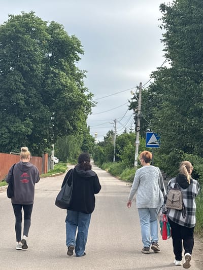 In the city of Sloboda, Russia, four individuals, viewed from the rear, walk down a paved road lined with trees and greenery. The sky is overcast. A red wooden fence runs along the left side of the road, followed by a patch of grass and then more trees. On the right side of the road, a utility pole with electrical wires and two streetlights is visible, along with a blue triangular traffic sign depicting a speed bump. Further down the road, a distant church with golden domes is visible. The first person, on the far left, wears a dark gray hoodie with red text and black leggings, carrying a black bag over their left shoulder and white sneakers. The second person wears a black hoodie and blue jeans, carrying a black bag. The third person is dressed in a light gray cardigan, light blue jeans, and brown shoes. The fourth person, on the far right, wears a black and white plaid hooded jacket, black pants, and white sneakers, carrying a backpack and a red and green bag.