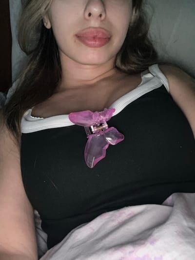 A close-up, overhead shot shows a person lying down on a bed, with their torso filling most of the frame. The person is wearing a black tank top with a white trim around the neckline and armholes. A translucent pink butterfly-shaped hair clip is attached to the front of the tank top, near the collarbone. The person's lips are slightly parted and appear to have a subtle shimmer. Their hair is a mix of brown and blonde and falls loosely around their shoulders. The background consists of a white wall and a pink and white patterned blanket. There is no visible indication of the city of Ust-Chorna, Ukraine, in this image.