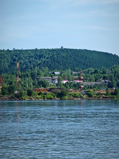 This is a wide, horizontal, eye-level shot of a settlement on the bank of a wide river. The sky is a pale blue with no clouds. The river in the foreground is a deep blue with ripples. The far bank of the river is lined with a dense, green forest that rises to form a rounded hill. In the middle ground, nestled among the trees, is a settlement with several buildings, some of which have red roofs. Several industrial structures are visible, including a tall, rusty yellow and brown pylon-like tower on the left and a large, red crane in the center. Piles of logs are stacked along the riverbank. The settlement appears to be a logging or industrial area. The image is undated and shows no people.