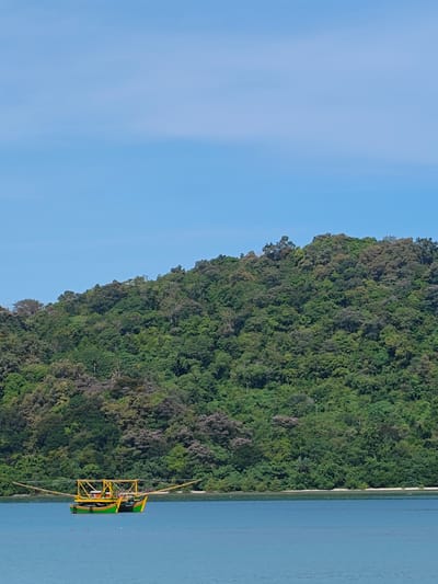 The image depicts a traditional Indonesian fishing boat in the water off the coast of Layeun, Indonesia. The boat has a distinctive configuration, with a green and yellow hull and outrigger arms extending from either side. It is located near a densely vegetated island with a blue sky above. The water is calm and blue, reflecting the sky.