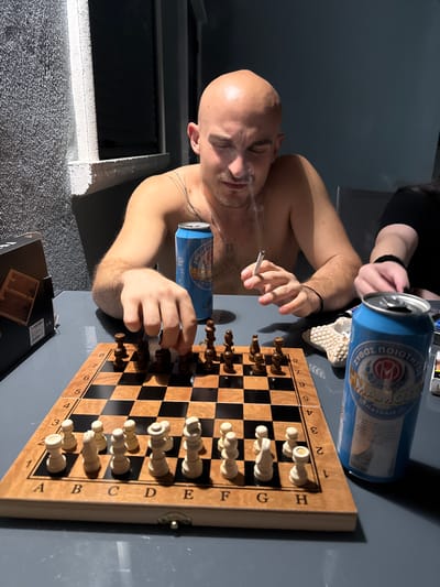 A man with a shaved head is playing chess at a table. He is shirtless and has a cigarette in his hand, from which smoke is rising. He is wearing a silver chain around his neck. On the table, there is a chessboard with pieces set up, two blue beer cans, and a box. A second person's hand is visible on the right side of the frame. The scene appears to be outdoors, possibly on a balcony or patio, with a wall and windows in the background. The lighting suggests it is evening or night.