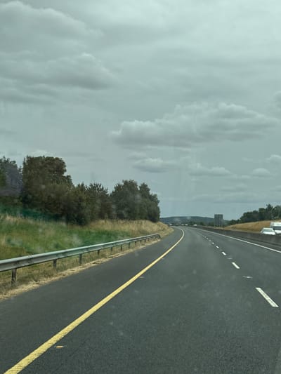 The image depicts a highway view with a cloudy sky. The road has a yellow line marking the left lane and white dashes marking the right lane. To the left of the road is a metal guardrail and a grassy embankment with trees. To the right of the road is a concrete barrier and more grassy embankments with trees in the distance. Several cars are visible on the highway, and a white van and another vehicle are parked on the right side of the road. The scene appears to be on a highway in the vicinity of Ballitore, Ireland, given the presence of vehicles and highway infrastructure. The lighting suggests it is daytime, but the overcast conditions cast a muted light over the scene.