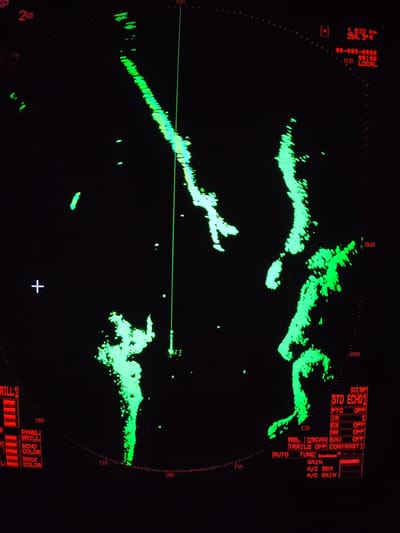 The image displays a marine radar screen with a green-on-black color scheme, showing detected echoes and navigational data. The central point of the screen, typically the ship's position, is not explicitly marked with a target, but a thin green line extends vertically from it, indicating the vessel's heading. The radar range is set to "2km", as indicated in the top-left corner, with fainter concentric dashed rings possibly marking smaller range increments.
Large, irregular bright green shapes are visible across the screen, primarily on the right and extending from the top-left to the bottom, likely representing landmasses or significant structures. Some smaller, isolated green specks also appear, which could be smaller vessels or clutter. A white cross mark is present in the lower-left quadrant, possibly a cursor or reference point.
Various operational and display settings are visible:
* **Top-left:** "SP" (possibly speed or ship position) and the "2km" range setting.
* **Top-right:** "1.610 km" and "294.3°R", indicating the range and bearing to a specific target or cursor position. Below this, " **-**-****-****", "**:**", and "(S) LOCAL" appear, likely for target tracking information or time/date.
* **Bottom-left control panel:** Labels include "DRILL 1", "PANEL", "BRILL" (brightness), "ECHO COLOR", and "BACK COLOR", with red bars next to them indicating current settings.
* **Bottom-right control panel:** Features radar display settings under "DISP STD ECHO1" such as "FTC OFF" (Fast Time Constant), "IR 1" (Interference Rejection), "ES OFF" (Echo Stretch), "NR OFF" (Noise Reduction), "REL |2SCAN| EAV OFF", and "TRAILS OFF CONTRAST 1". Below these, "AUTO TUNE", "GAIN", "A/C SEA", and "A/C RAIN" are visible, likely for automatic tuning and clutter suppression settings.
* **Bearing markings:** Radial lines and numbers around the perimeter indicate bearings, such as "060", "090", "120", "150", "180", "210", and "240".
The image captures the active state of a radar system, likely being used for navigation or monitoring the surrounding environment at sea. There are no direct cues in the image to indicate time of day or specific weather conditions beyond the radar's clutter suppression settings. The location context of Topornya, Russia, is not identifiable from the radar display itself.
