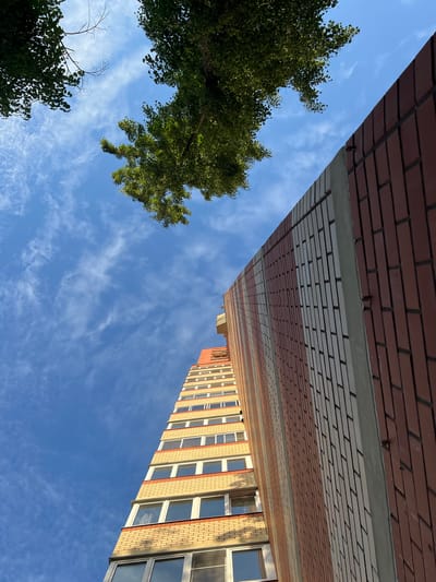 The image depicts a low-angle view looking up at a tall, modern apartment building against a bright blue sky with wispy white clouds. The building is constructed with alternating bands of light yellow and red brick, interspersed with rows of windows. A portion of a nearby brick wall, facing the apartment building, is also visible on the right side of the frame. Green foliage from trees frames the top left of the image. The scene appears to be in a contemporary urban or suburban setting.