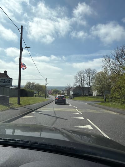 The image, taken from inside a vehicle, shows a street in Witton Gilbert, United Kingdom. The view is from the driver's seat, looking down the road. A car is visible ahead on the road. Road markings, including white triangles pointing inwards, are present. On the left side of the road, a sign for Witton Gilbert is affixed to a utility pole, which is also decorated with a Union Jack flag. The sky is visible with scattered clouds. Trees and vegetation line the roadside.