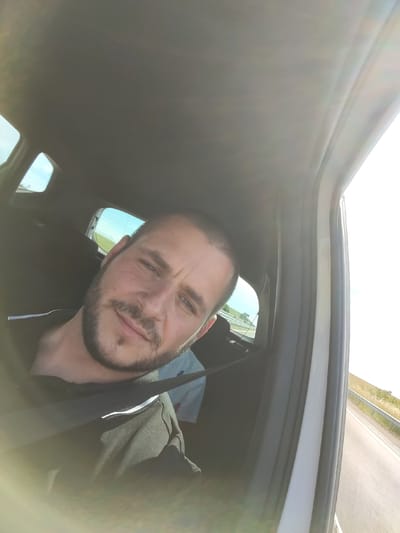 The image is a selfie taken from inside a car, possibly on a highway, with a blurred view of the surrounding landscape, which appears to be rural. The main subject is a man with short brown hair and a beard, wearing a black polo shirt. He is looking towards the camera with a slight smile and a seatbelt is visible across his chest. The car's interior is visible, including the roof lining and windows. In the background, through the rear window, a second person is partially visible, wearing a light-colored t-shirt. The lighting suggests it is daytime, with sunlight streaming into the car. The location is identified as the city of Troyanovo, Bulgaria.