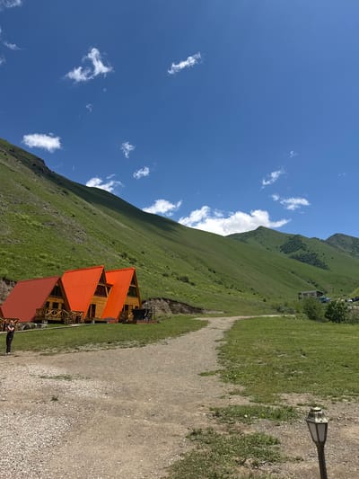 The image shows a scenic view of rolling green hills under a clear blue sky with scattered white clouds. In the foreground, there are three A-frame wooden cabins with orange and red metal roofs. A gravel path leads towards the cabins and continues into the distance, winding up into the hills. A person is visible on the left side of the image, standing near the cabins and holding a phone, seemingly taking a photo. A decorative lamppost stands on the right side of the path. In the background, the lush green slopes of the mountains are prominent, with a few sparse trees and shrubs dotting the landscape. The overall scene depicts a serene, natural environment, likely a rural or mountainous area. The location is identified as El'tiubiu, Russia.