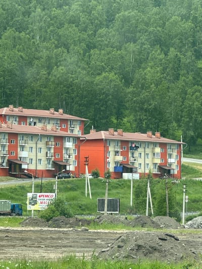 The image depicts a residential area in Slyudyanka, Russia, with multi-story buildings set against a backdrop of a dense forest. Two apartment buildings, painted in a combination of yellow and orange, are prominently featured. These buildings have red-tiled roofs with visible chimneys and multiple balconies with railings. Several windows are visible on the facades of the buildings.
In front of the buildings, a street is visible, with a parked black car and a few people standing near it. A blue truck with a cargo container is parked on the left side of the image. A billboard with Russian text and images is also present, advertising "PEMCO".
The environment consists of grassy slopes, some areas with piles of dirt or gravel, and utility poles with wires. The overall impression is a functional, somewhat utilitarian residential neighborhood bordering a natural wooded area. The image appears to be taken during daylight, with overcast conditions contributing to a muted color palette.