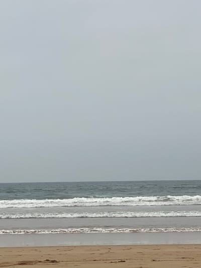 The image shows a view of the ocean, likely taken from indoors. The sky is overcast and a pale gray color. The ocean is a darker gray, with white foamy waves visible. A wooden frame is visible on the right side of the image. The location is likely Taghazout, Morocco, given the mention of the city. No people, specific objects, or actions are discernible beyond the ocean and waves.