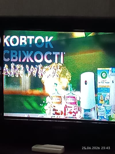 This image shows a television screen displaying text and product packaging against a green background. The text in white reads "КОВТОК" and below it, "СВІЖОСТІ". Underneath this, there is more text which is partially obscured and appears to read "AIR WAVES".

On the right side of the screen, there are several product packaging items, including a white cylindrical object with a dark circle and a blue box with a green logo. In the foreground, on the bottom right of the image, a timestamp reads "25.06.2026 23:43".

The image is taken indoors, likely in a home, with the television placed on a surface. The overall scene is blurry and has some digital distortion, characteristic of a photograph taken of a screen. There is no identifiable location in the city of Bolhrad, Ukraine, visible in this image.