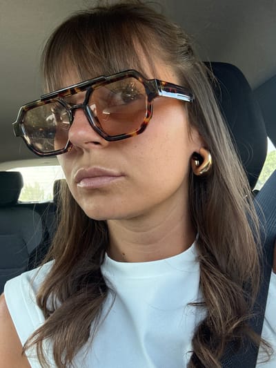 The image features a close-up portrait of a person, rotated 90 degrees clockwise, appearing to be taken inside a vehicle in Le Muy, France. The subject has long, wavy brown hair, which falls over their shoulders and chest. They are wearing a simple white t-shirt or top.

A prominent feature is a pair of oversized eyeglasses with thick, angular, tortoiseshell-patterned frames. The lenses are tinted, and reflections within them show glimpses of the vehicle's interior and some outdoor foliage, suggesting it is daytime. The person is looking slightly downward and to the left, through these glasses, towards the camera.

On the visible ear, a distinctive, polished gold earring or ear cuff with an abstract, sculptural design is worn. The person's lips are a natural tone, possibly with a subtle sheen. The background is mostly dark and out of focus, revealing hints of dark upholstery typical of a car seat. No discernible text is visible in the image. The overall impression is a casual, relaxed moment captured while traveling or seated in a car.