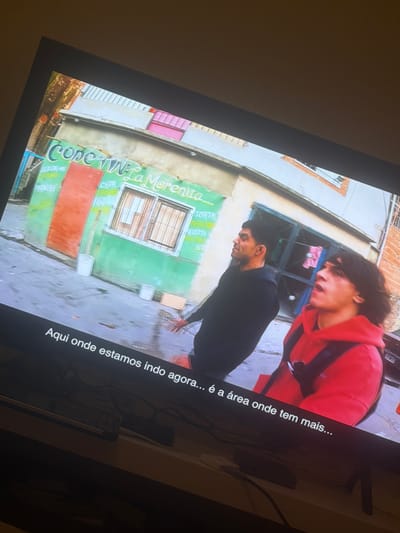 The image shows a scene from a TV screen, displaying a building with the name "Cope Wi La Morenita" painted on it. The building is painted green with a red door. The text below the sign lists items such as "CHIPA," "SANDH," and "TORTAN." Two men are walking in front of the building, one wearing a black outfit and the other a red hoodie. There is text overlayed on the screen in Portuguese, reading "Aqui onde estamos indo agora... é a área onde tem mais..."