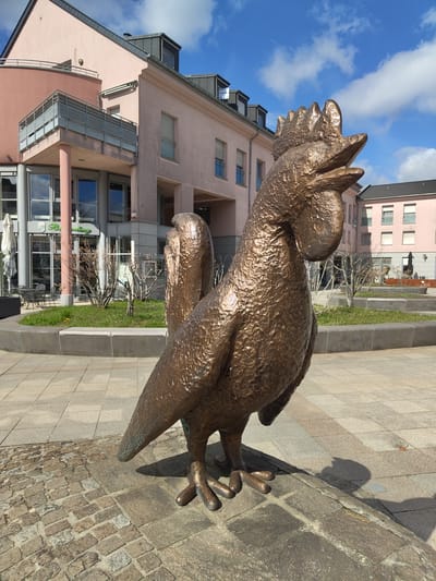 A textured bronze statue of a rooster stands prominently on a paved public square in Bertrange, Luxembourg. The statue, facing right with its beak open, displays a detailed comb and wattle. It rests on an area of irregular cobblestones bordered by light grey square pavers.

In the mid-ground, a landscaped section features raised dark grey concrete planters containing sparse green grass and several bare deciduous shrubs. Beyond this, a multi-story building with a light peach-colored stucco facade and dark grey roofline dominates the left and central background. This building has a glass-fronted ground floor, revealing an interior, with an exterior sign partially displaying "Bertrange." An outdoor patio area equipped with white parasols, dark tables, and chairs is visible adjacent to the building. Another building of similar architectural style and color is visible further to the right. The sky overhead is blue with scattered white cumulus clouds, and direct sunlight illuminates the scene, casting distinct shadows. No individuals are present.