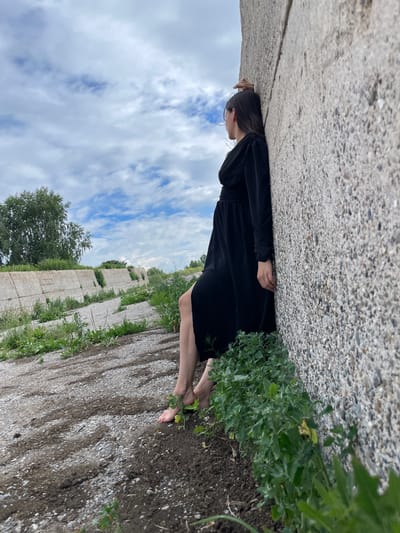 A young woman in a black dress is lying down outdoors, resting her head on a concrete wall. She is barefoot and appears to be relaxed or contemplative. The setting is a textured, grey concrete surface meeting a patch of green foliage. Beyond this, a long, low concrete structure is visible, with vegetation growing alongside it. In the background, trees and a cloudy sky with patches of blue are seen. The lighting suggests it is daytime, possibly overcast or with diffused sunlight, given the soft shadows. The overall mood is one of quiet solitude and connection with the natural environment, despite the man-made elements of the scene. No visible text is present in the image.