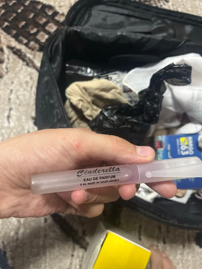 The image shows a person holding a small, translucent, cylindrical bottle of perfume. The bottle has a white cap and a pale pink liquid inside. The text on the bottle reads "Cinderella EAU DE PARFUM 8 ML MADE IN SAUDI ARABIA". In the background, a black open bag is visible, with some white and black plastic objects inside, and a beige piece of fabric. There are also parts of a yellow box and a blue and yellow poster with text and an image of a bus. The scene appears to be indoors, possibly in a home, and the lighting suggests daytime. The person's hand is visible holding the perfume bottle. The image is cropped to focus on the perfume bottle and the person's hand, with the background elements blurred.