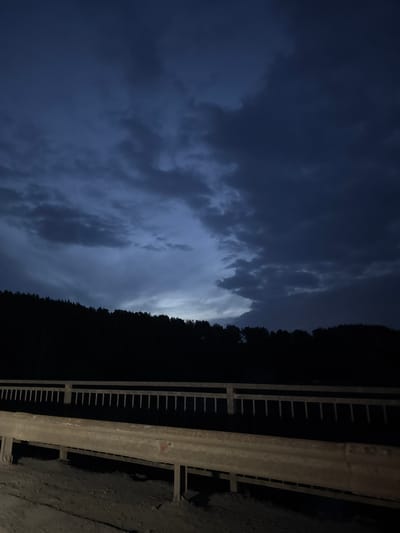 The image depicts a night scene, likely taken from a bridge or elevated roadway in Doksha, Russia. The left side of the image showcases a cloudy, dark blue sky with visible cloud formations. To the right, a dense, dark forest or line of trees is visible. In the foreground, a gray metal guardrail runs along the right side of the frame. The scene is dimly lit, suggesting either a lack of streetlights or nighttime conditions.