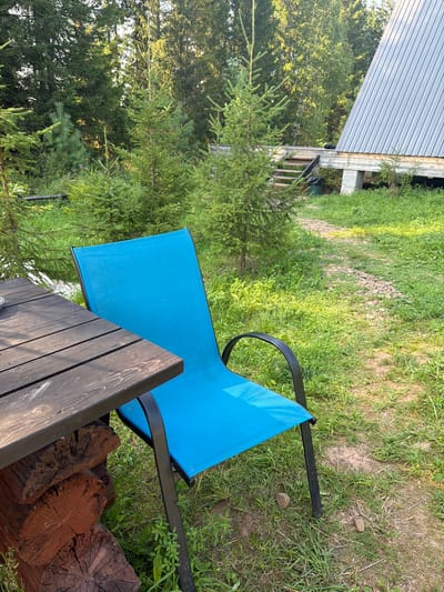 The image depicts an outdoor scene in the city of Kuliki, Russia, featuring a bright blue chair positioned next to a dark wooden table. The table is supported by rough-hewn logs. The ground is covered in lush green grass, with a gravel path visible in the background. A modern, A-frame style building with a corrugated metal roof is situated to the right, accessible by a wooden staircase. Evergreen trees, likely firs, surround the area, creating a natural, wooded ambiance. The lighting suggests it is daytime, with sunlight filtering through the trees, casting shadows on the grass. There are no people visible in the image.