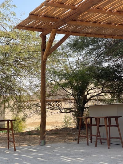 The image displays an outdoor patio area situated within the Arava Tichona Regional Council, Israel, under a clear, bright sky. A rustic pergola, constructed from raw wooden logs and branches, extends across the upper and left sections of the frame, casting distinct dappled shadows onto the light-colored, compacted ground. A primary vertical wooden support post for the pergola is prominent on the left, secured at its base with a metal band and diagonally braced to an overhead beam. To the right, a light-toned, unstaffed service counter is visible, with a metal faucet installed behind it. Two unoccupied wooden bar stools, characterized by contoured seats and splayed legs, are positioned in front of the counter. The mid-ground features dry, reddish-brown earth interspersed with abundant green foliage from various trees, including fine-leafed species and a larger, darker-leafed tree. In the background, partially obscured by vegetation, is a light-colored building constructed of concrete blocks, exhibiting a decorative or ventilation pattern along its upper section.