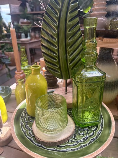 A tabletop display of green glass objects is presented. In the foreground, a green, intricately carved plate rests on a wooden tray. On the plate are two green glass items: a cylindrical glass with a textured pattern, supported by a slice of wood, and a tall, square-shaped bottle with decorative relief. To the left of these items, a smaller, bulbous green bottle is visible. Behind these, a tall, slender green glass bottle stands. Further back, a large, green, leaf-shaped decorative object is prominent. A cream-colored candle on a wooden base is to the left of the leaf. The background is blurred, showing shelves with various decorative items, including ceramic vases and plants, suggesting an indoor retail or decorative setting. The scene is indoors, possibly in Fritzlar, Germany, given the prompt. No people are present in the image.