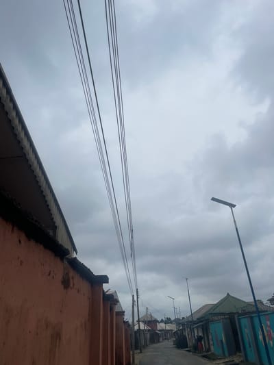 In Iwofe, Nigeria, a street scene unfolds under a heavily overcast sky. On the left, a textured, reddish-orange wall runs along the foreground, topped by a corrugated metal roofline with a decorative, scalloped edge. Overhead, a series of thick black electrical cables stretch across the frame. The street itself is paved and extends into the distance, lined with various buildings. Some structures feature green metal roofs and walls adorned with colorful cartoon-like murals. Several streetlights are visible along the road, including one with a blue pole. A few figures are present on the street, one a person in a reddish garment standing near an entrance, and another further down the road appearing to walk away from the camera. The overall impression is of a quiet, everyday street in a Nigerian town on a cloudy day.
