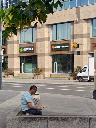 A man in a light blue t-shirt and blue jeans sits on a concrete bench, looking at his phone. He is on a sidewalk in front of a modern building with a beige and gray stone facade. The building has large windows, and signs for "МИРАТОРГ" (MIRATORG) and "ВКУСНО-И-ТОЧКА" (VKUSNO-I-TOCHKA) are visible. A white delivery truck is parked on the street to the right of the building. Trees with green leaves and yellow flowers are hanging over the scene from the upper left. The scene is set in an urban environment, likely a commercial district in Moscow, Russia, given the Cyrillic signage.