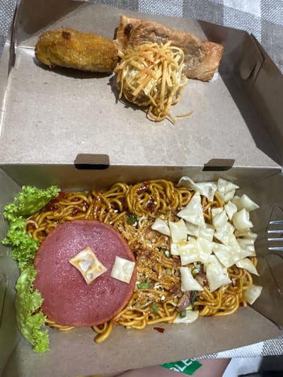 The image shows a close-up, overhead view of a takeout box filled with food. The box is made of cardboard and is positioned on a surface with a checkered pattern, possibly a tablecloth.

In the upper section of the box, there are three distinct food items. On the left is a fried, breaded cylindrical item, likely a croquette or spring roll. In the center, there is a mound of shredded, fried noodles topped with a lighter colored shredded ingredient, possibly cheese or fried shallots. To the right of this is a rectangular piece of fried food, possibly tofu or a cutlet.

The lower section of the box contains a generous portion of stir-fried noodles. The noodles are coated in a reddish-brown sauce and mixed with various ingredients, including what appears to be chopped green onions and crispy fried bits. There is a large, round slice of pink ham or mortadella placed on top of the noodles on the left side, adorned with two small, square pieces of fried wonton wrapper. On the right side of the noodles, there are several irregularly shaped, light-colored, crispy pieces, likely fried wonton wrappers, scattered across the dish. A few sprigs of bright green lettuce are visible on the far left of this section. A silver fork is partially visible on the right edge of the box.

The setting is identified as Kalijaten, Indonesia. The image captures a meal presented in a disposable takeout container.