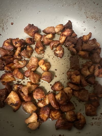 The image presents a close-up, overhead view of numerous small, diced pieces of meat, likely pork belly or a similar fatty cut, being cooked in a pan. The meat pieces are varied in color, ranging from a golden brown to a rich, dark brown, indicating different stages of browning and caramelization. Some pieces appear crispy and well-rendered, with a textured surface, while others show shinier, more translucent areas of rendered fat.
The meat is distributed across the bottom of a light-colored pan, which appears to be made of metal with a non-stick or well-worn surface. Around and beneath the meat, there are visible pools of rendered fat or oil. The pan surface also shows small, dark, irregularly shaped specks and residues, consistent with food particles that have browned or stuck during cooking. Small droplets of liquid (likely fat or moisture) are scattered across the empty areas of the pan.
The scene captures an activity of food preparation, specifically the frying or rendering of meat. The focus is entirely on the food and cooking vessel, with no people visible. The setting is an indoor kitchen. There is no visible text in the image.