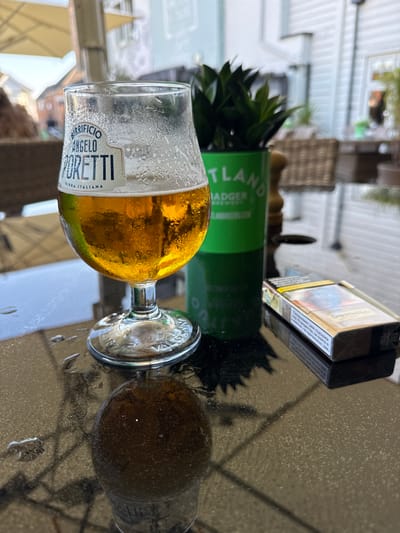 A close-up, eye-level shot captures a glass of golden beer with condensation on a wet, reflective table, likely outdoors at a cafe in Poole, United Kingdom. The glass is a branded "Birrificio Angelo Poretti" with "Birra Italiana" printed on it. To the right of the beer glass, a pack of Dunhill cigarettes rests on the table, partially obscuring a green pot containing a dark green plant. The background is blurred, showing hints of outdoor seating, umbrellas, and building exteriors, consistent with a cafe or restaurant environment. Water droplets are visible on the dark table surface, suggesting a recent rain shower or a damp environment.