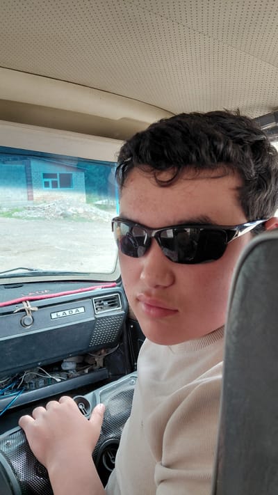 The image shows a person, likely a young male, sitting in the driver's seat of a vehicle, possibly a Lada based on the visible text. He is wearing sunglasses and a light-colored, long-sleeved shirt. The interior of the vehicle is visible, including the dashboard with a speaker and what appears to be engine components. The person's right hand is resting on the dashboard. Through the windshield, a building with a blue-tinted window and what looks like a rural landscape are visible. The sun appears to be shining, suggesting it's daytime. The overall setting appears to be a road or a parked location in a rural area. The person's expression is neutral, and they are looking towards the camera. The image is oriented sideways.