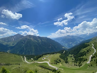 The image captures a panoramic view of a mountainous landscape under a bright blue sky dotted with scattered white clouds. A winding paved road snakes through lush green rolling hills and dense evergreen forests. Several small buildings, possibly chalets or farmhouses, are visible nestled on the slopes. In the distance, towering mountains with some snow-capped peaks dominate the horizon, with a valley stretching out below. The scene suggests a scenic location in the Alps, likely during a sunny day in the warmer months. The location is identified as St. Leonhard in Passeier - San Leonardo in Passiria, Italy, a town known for its picturesque alpine surroundings. There are no people or animals visible in the frame, and the road appears to be either empty or the vehicles are too small to discern.