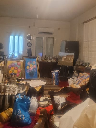 The scene depicts a highly cluttered interior room, consistent with a residential space that also functions as an artist's studio. The specific city of Κωστήτσι, Greece, as provided, cannot be independently verified from the image alone. No individuals are present.
The room is illuminated by interior lighting, with a window on the left wall featuring light-colored curtains, possibly sheer. An air conditioning unit is mounted high on the left wall. Below the window, a round table draped with a white crocheted tablecloth holds art supplies, including paintbrushes in a glass jar, a small metallic pot, and various small containers. Two framed paintings are prominently displayed upright on or next to this table. The left painting, in a gold-colored frame, depicts a bouquet of multicolored flowers (pinks, purples, yellows, blues) against a yellow background. The right painting, in a darker frame, features a similar bouquet of colorful flowers against a bright blue background.
On the floor to the left, a striped cylindrical pillow rests alongside a large blue fabric shopping bag with a white "AB" logo and a white plastic bag. In the central area, a light-colored easel holds a framed landscape painting depicting a stone building under a pale sky. Behind the easel, a dark wooden cabinet is visible, topped with assorted bottles and items. A tall, dark-colored refrigerator or cabinet with magnets is positioned on the right side of the room. Adjacent to it, a long wooden table is heavily laden with objects, including a vintage sewing machine, textiles, and papers.
The floor throughout the room is covered with various items, including additional plastic bags, textiles, pillows (one red, one checkered), and what appears to be clothing. A dark-colored, possibly electric, heater or fan unit is visible in the foreground right. Partially obscured in the foreground right is a wooden armchair with carved details. The far right wall displays a textured pattern, possibly wallpaper. The overall environment suggests a lived-in space with significant artistic activity and general storage.