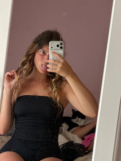 The image is a mirror selfie taken indoors, likely in a bedroom. The main subject is a young woman with long, wavy blonde hair. She is wearing a black, strapless, ruched garment. In the reflection, she is holding a white smartphone to take the photo, making a playful face with her tongue sticking out and a tongue piercing visible.

A black and white cat is visible in the background, lying on a pile of clothes. The setting appears to be a room with a pinkish-purple wall reflected in the mirror, and some light-colored bedding or clothing is also present. The overall mood is casual and lighthearted. The time of day is not discernible, but the indoor setting suggests it could be anytime. No visible text is present in the image. The location context of Sainte-Marie-la-Mer, France, does not provide specific visual cues within the image itself.