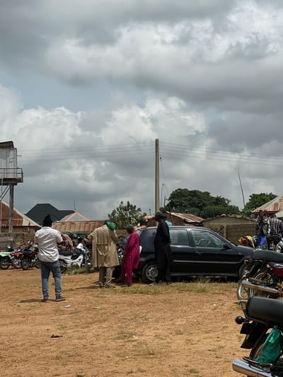 The image depicts an outdoor scene in Kassa, Nigeria, under a cloudy sky. Several people are gathered around a black sedan and a collection of motorcycles.

In the foreground, the ground is composed of reddish-brown dirt with sparse patches of dry grass. A man in a white striped shirt and blue jeans stands facing away from the camera, his back to a group of motorcycles. To his right, a man in a tan coat stands with his back to the camera, holding a green object in his left hand. A woman in a bright pink garment is to his right, facing the car. Another person in black attire is standing on the other side of the car.

The black sedan is parked in the middle ground, with its doors slightly ajar and its front wheels turned. A row of motorcycles is parked to the left of the sedan. Further to the left, a water tower structure is visible, with corrugated metal roofing on adjacent buildings.

In the background, more buildings with corrugated metal roofs are visible, interspersed with trees. A wooden utility pole with wires runs across the scene. The sky is filled with heavy, grey clouds, suggesting an overcast or potentially rainy day. The overall impression is one of a community gathering or a temporary stop in a semi-urban or rural area.
