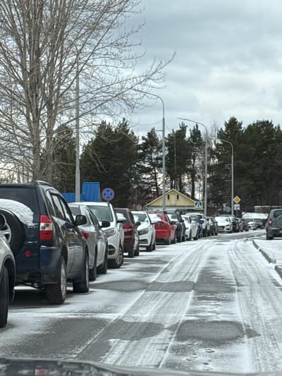 The image shows a line of cars parked on the side of a snowy road in Beloyarsky, Russia. The cars are parked close together, facing in the same direction. There are trees and buildings in the background. The sky is overcast. A bare tree is visible in the foreground. Several road signs are present along the road. The road is icy.