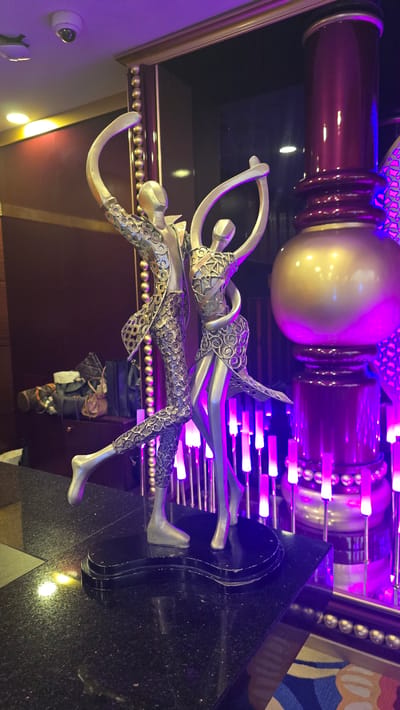 A golden sculpture of a dancing couple stands on a dark, polished surface. The male figure wears a jacket with an intricate pattern and patterned trousers, while the female figure has a patterned top and a skirt that appears to be made of flowing fabric. Both figures have their arms raised and are posed in a dynamic dance. Behind them, a large, glossy purple cylindrical object with a golden sphere on top is illuminated by vibrant purple lights. The background includes a dark, reflective surface, possibly a mirror, and a maroon-colored wall. To the left, there are several handbags and other objects on a shelf. The lighting is dramatic, with strong purple and yellow hues. The scene appears to be indoors, possibly in a hotel lobby or a similar public space, given the decorative elements and the presence of the security camera visible in the top left corner. The location is identified as Kazafani, Northern Cyprus / Cyprus.